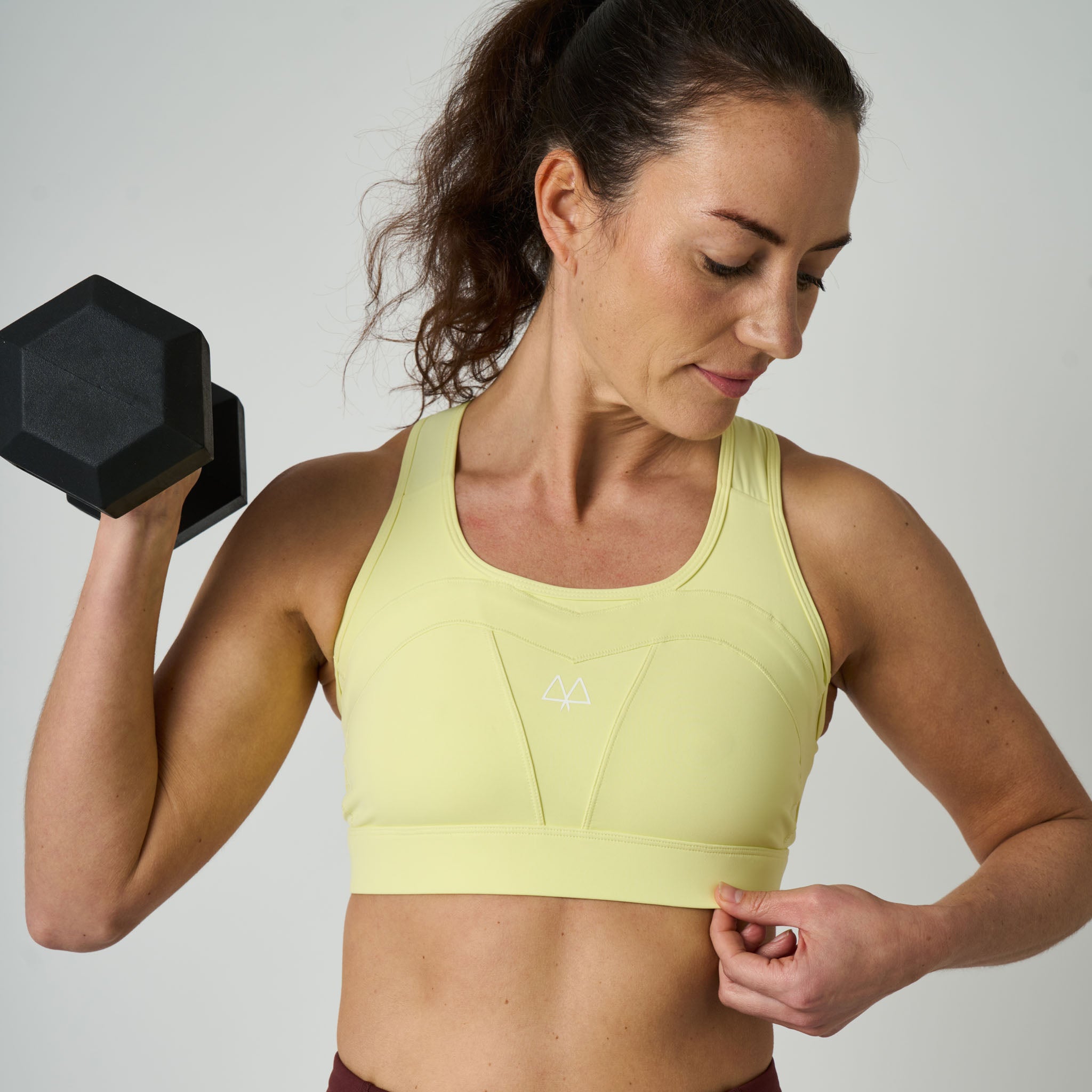 Woman in a yellow sports bra holding a black dumbbell against a plain background